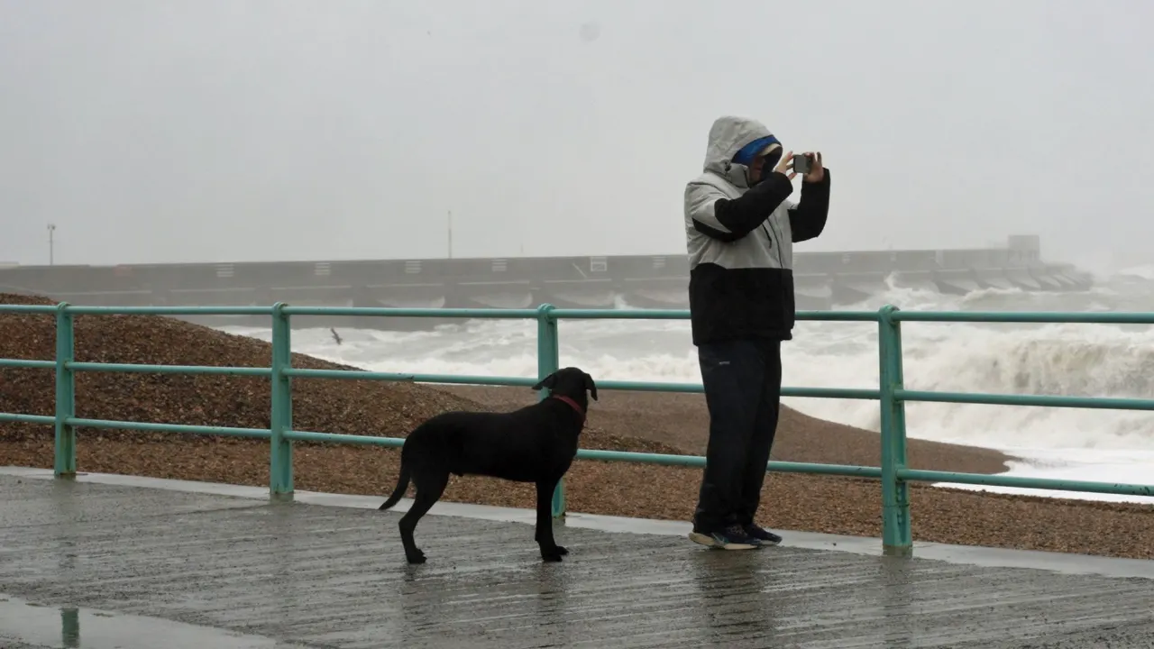 Hooded person photographs rough sea waves from railed seafront promenade beside a black dog during stormy weather.