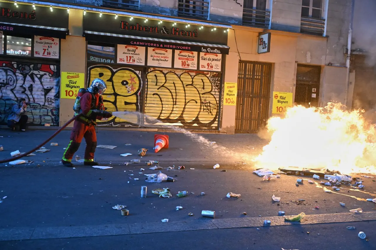Firefighter in red gear sprays fire hose near burning debris on littered Paris street at night