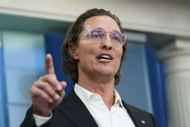 A man in glasses and dark blazer gestures with raised finger while speaking indoors.