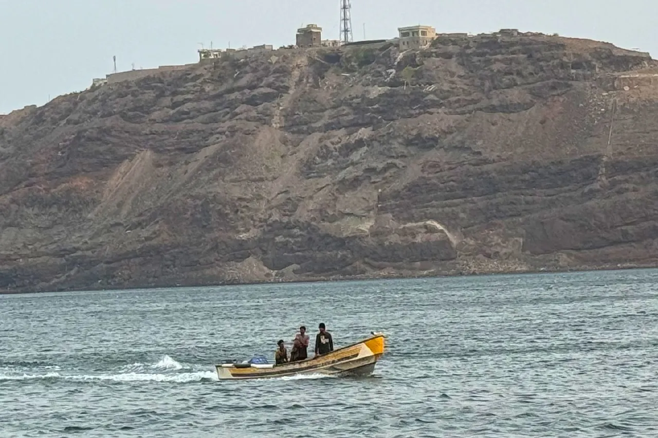 Small wooden boat with three men on calm sea before rocky cliff with buildings on top.