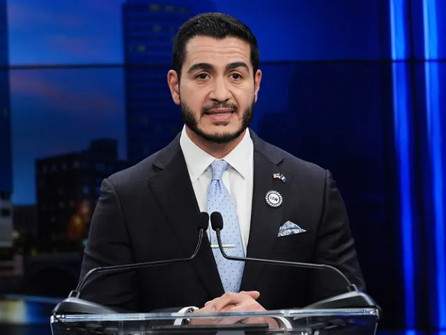 Man in dark suit and blue tie speaking at a clear podium before a blue studio backdrop.