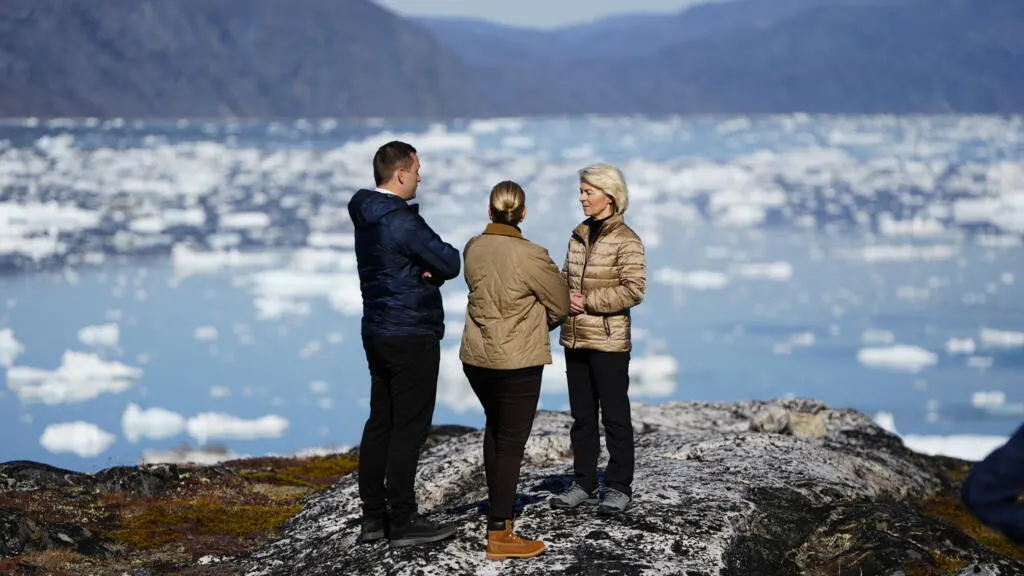 Three people talking on a rocky outcrop overlooking icebergs floating in a fjord.
