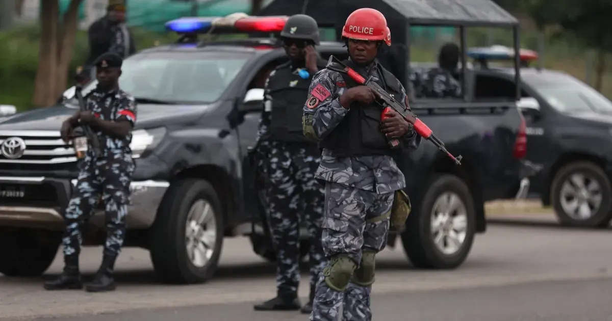 Armed police in camouflage uniforms with red helmet and rifle beside parked police vehicles on a street.
