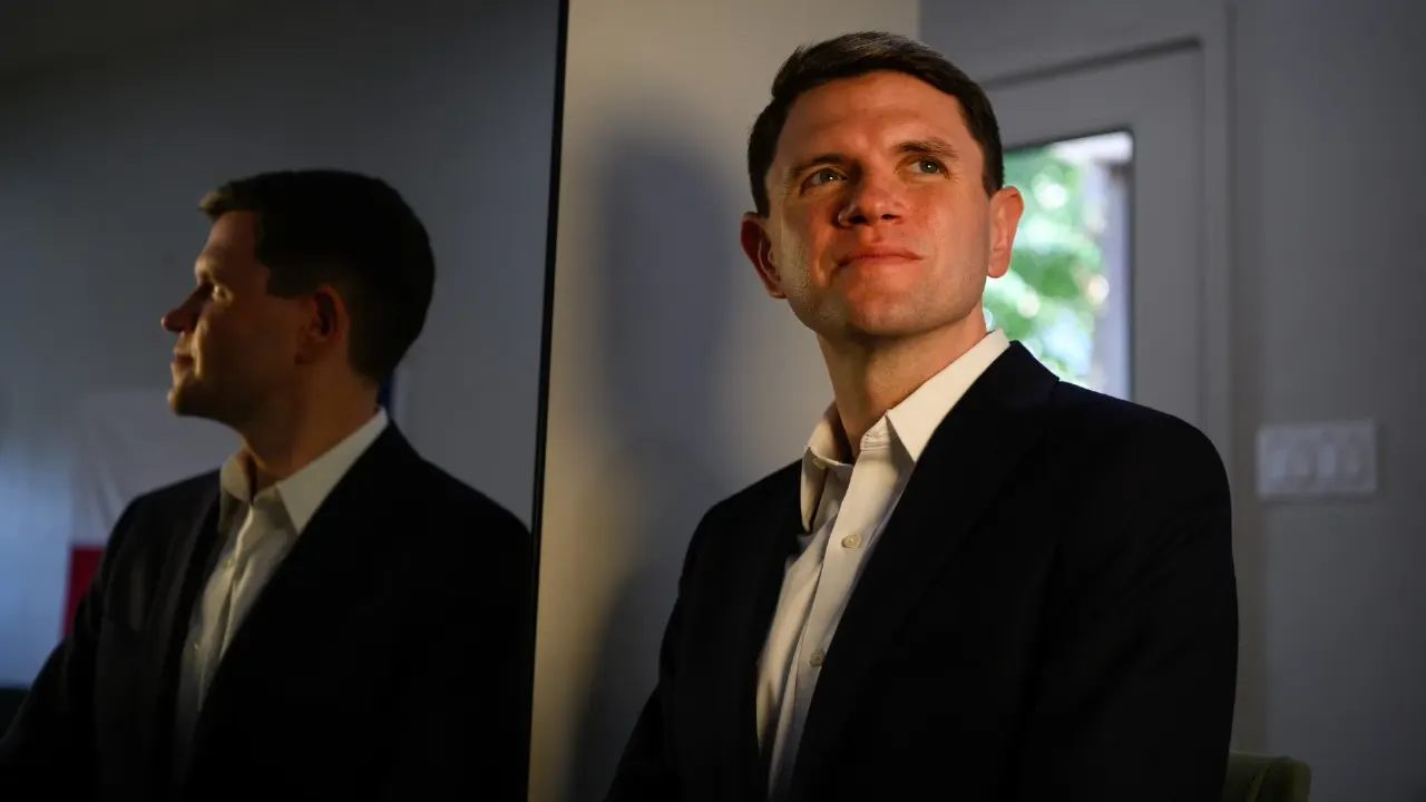 A man in a dark suit and white shirt gazes upward beside a reflective mirror in a room.