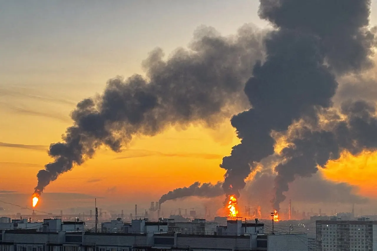 Thick black smoke and flares rising from an industrial refinery at sunrise over residential buildings.