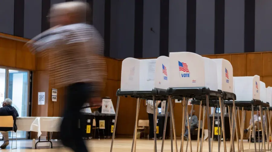 Blurred voter walks past rows of white voting booths with American flags and VOTE signs.