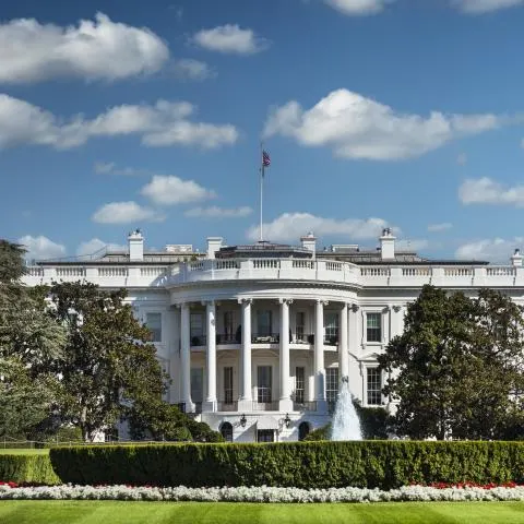 The White House south facade with columns, lawn, fountain, and flagpole under blue sky.