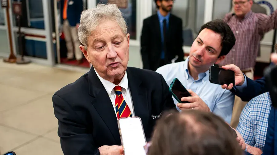 Older man in dark suit speaks to reporters holding phones in indoor hallway