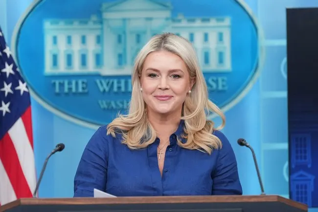 Smiling woman at a podium before White House seal backdrop, with American flag.