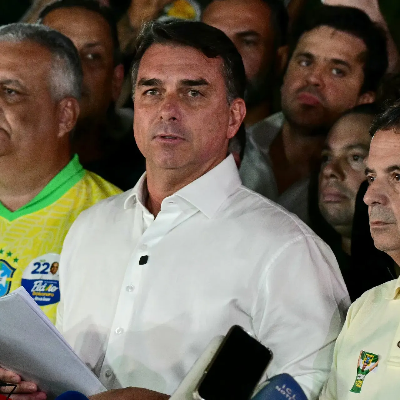 Man in white shirt speaking at microphones before a crowd of supporters in yellow campaign shirts.