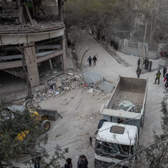 Aerial view of a bombed building's rubble on a street, with a dump truck and onlookers.