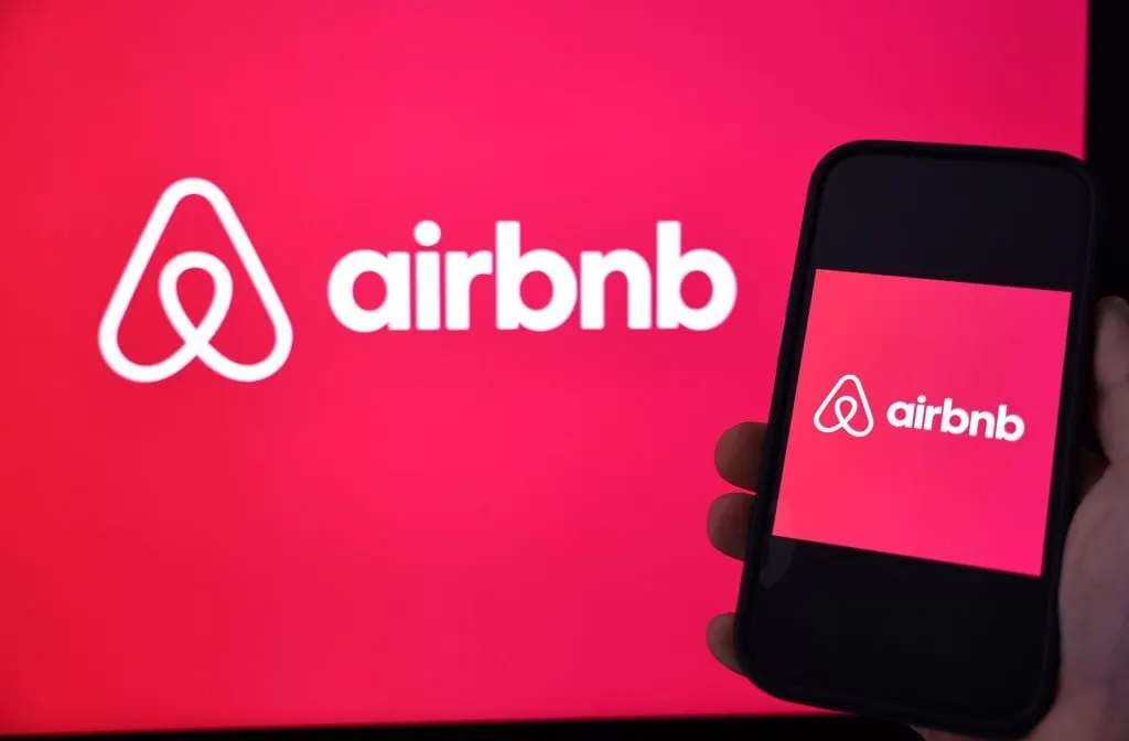 Airbnb logo on a large red screen beside a phone showing the same logo, held by a hand.