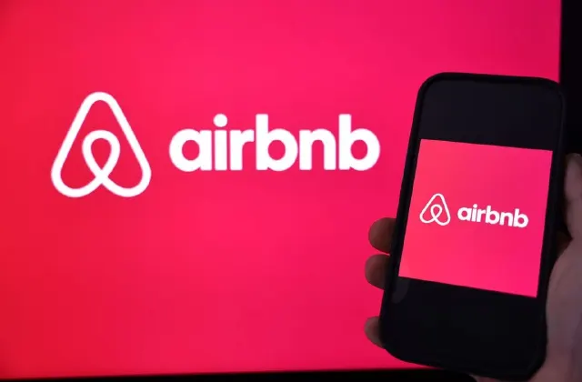 Airbnb logo on a large red screen beside a phone showing the same logo, held by a hand.