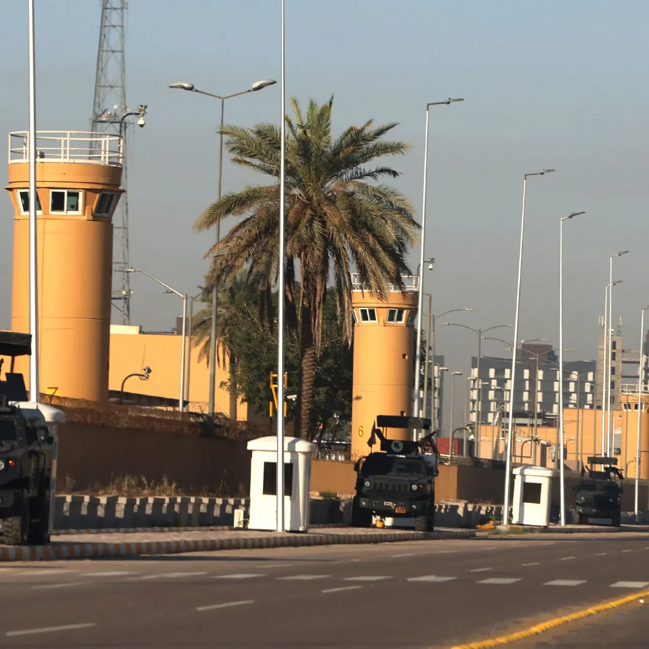 Armored security vehicles and guard towers lining a road at a fortified compound with palm trees