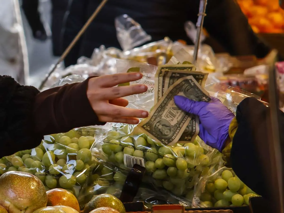 Gloved hand holding dollar bills over bagged grapes at a market stall, another hand reaching in.