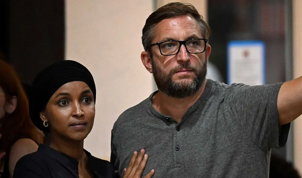 Two people standing indoors, a woman in a black head covering and a bearded man in glasses pointing.