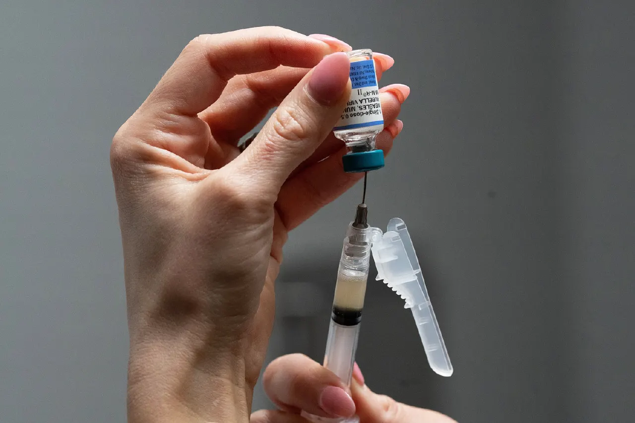 Gloved hands filling a syringe from a vaccine vial against a plain grey background.