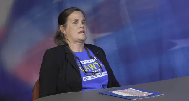 Woman in black blazer and blue campaign shirt seated at desk, mid-speech, against blue backdrop