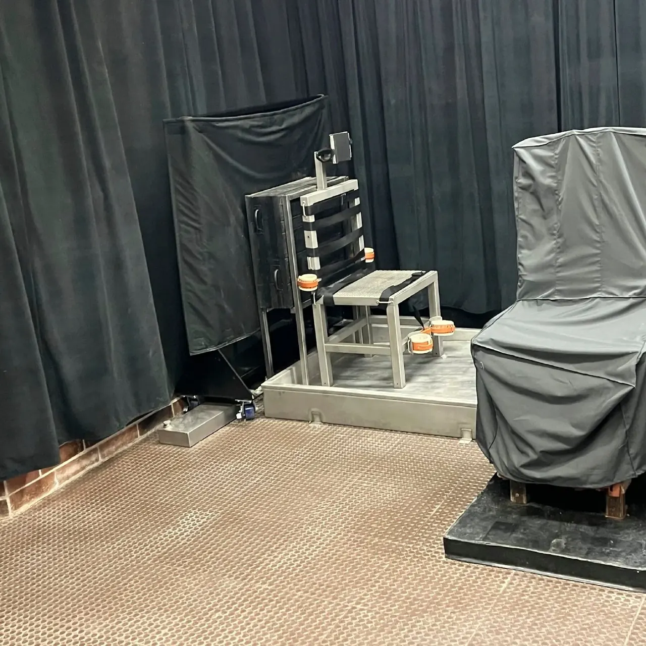Execution chamber with chair apparatus and medical equipment behind dark curtains.