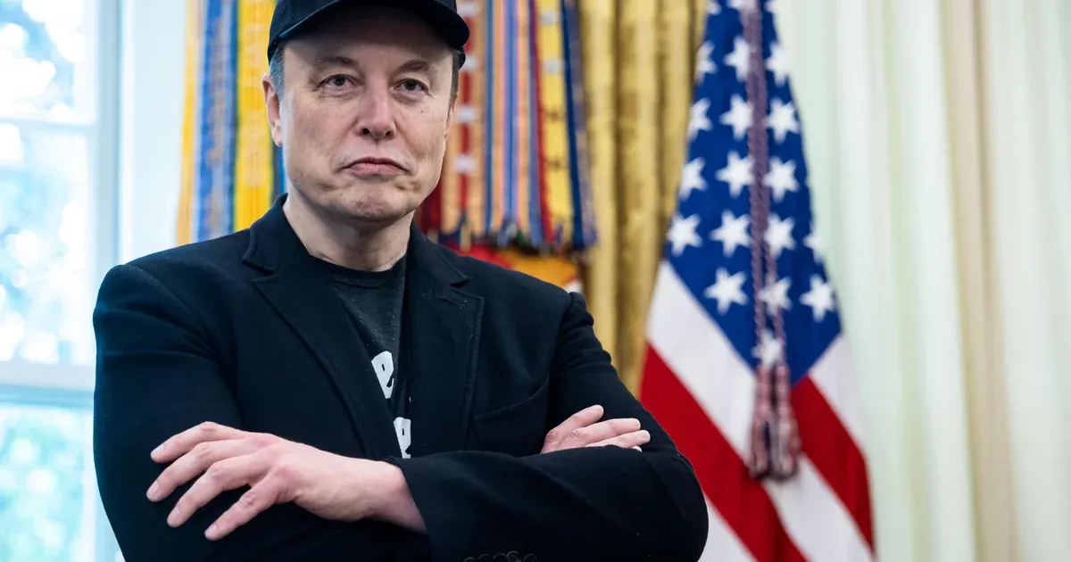 Man in black cap and jacket with arms crossed, standing before an American flag and ribbons.