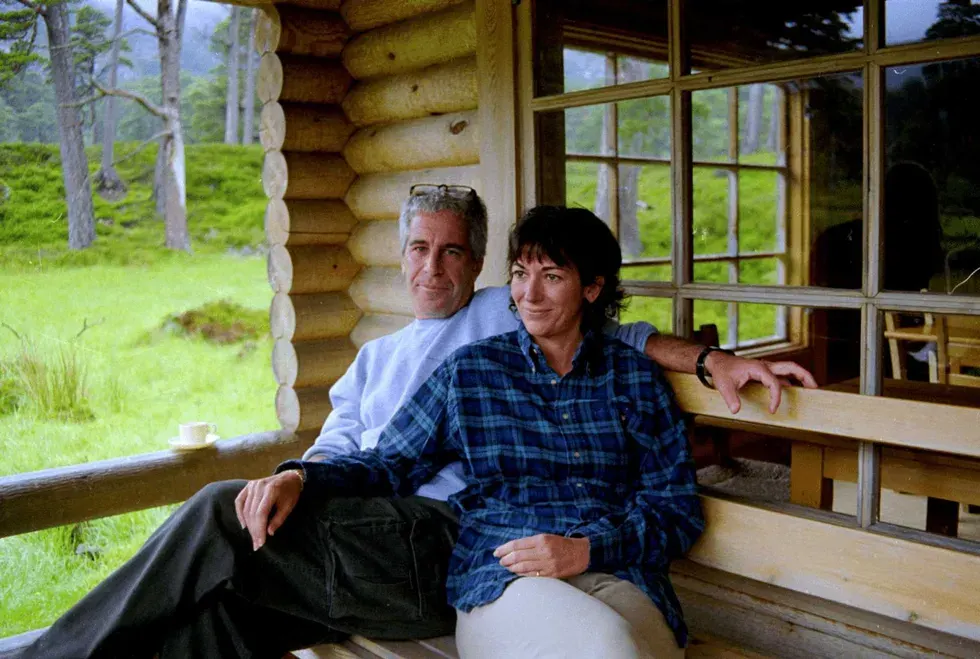 Two people sit together on a log cabin porch overlooking green hillside and trees.