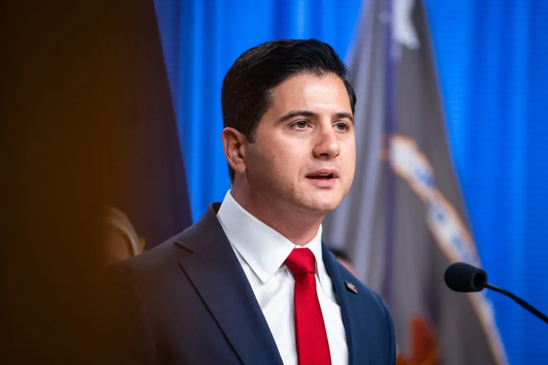 Man in dark suit and red tie speaking at a press microphone before blue curtains and flag