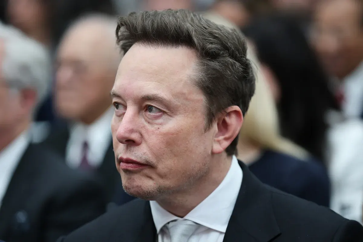 Man in dark suit and white shirt looking sideways at a blurred crowd indoors.