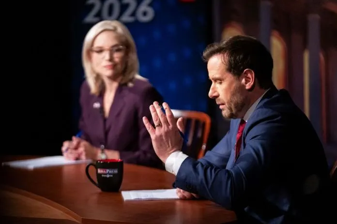Two debate candidates seated at a round table, one gesturing mid-speech, with an Iowa PBS mug.
