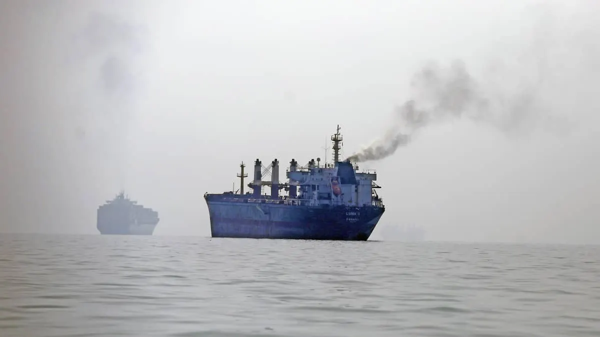 Blue cargo ship emitting smoke at sea in hazy fog, with another vessel behind it.