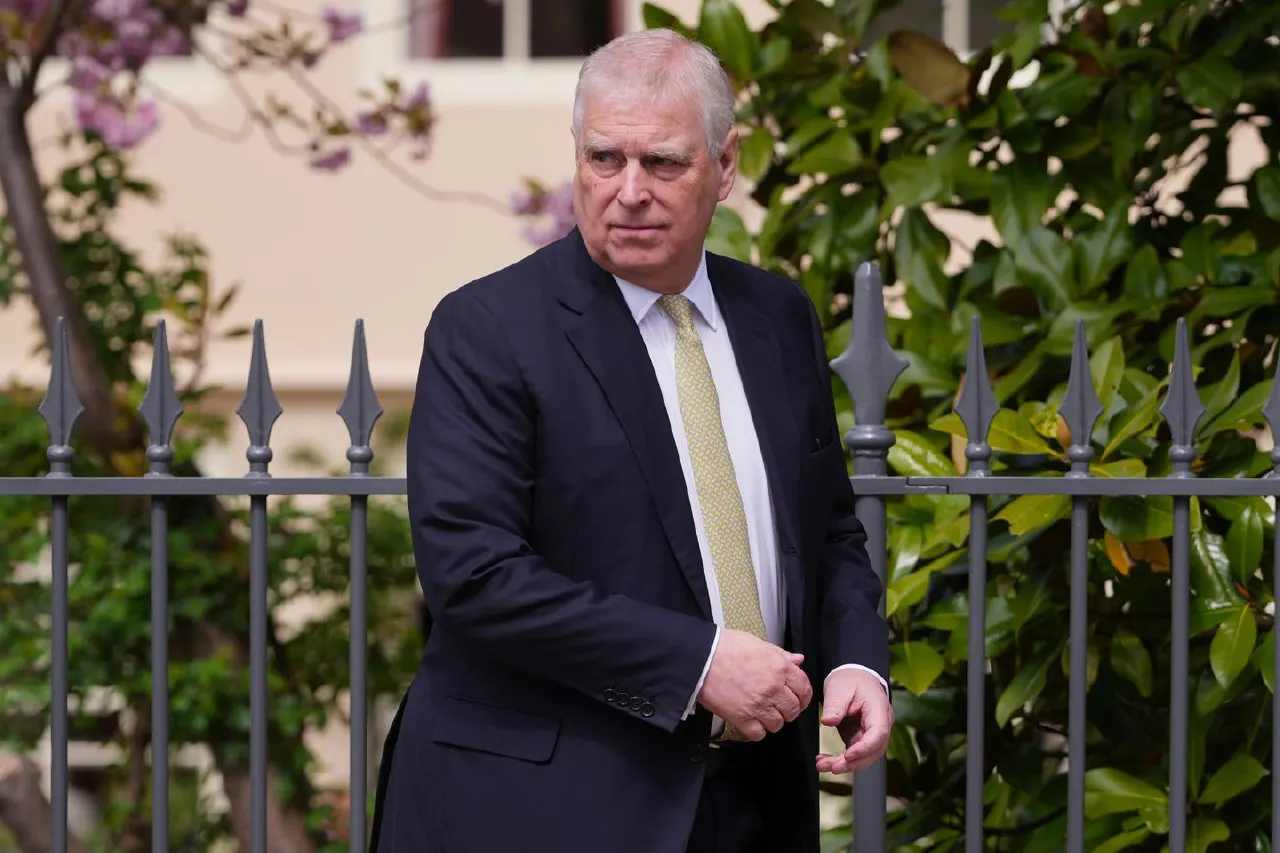 Man in dark suit and yellow tie standing behind black iron railings, greenery behind him.