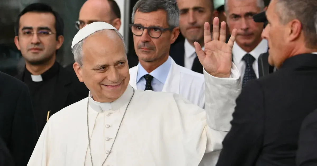 Elderly man in white papal-style vestments smiling and waving, surrounded by clergy and staff.