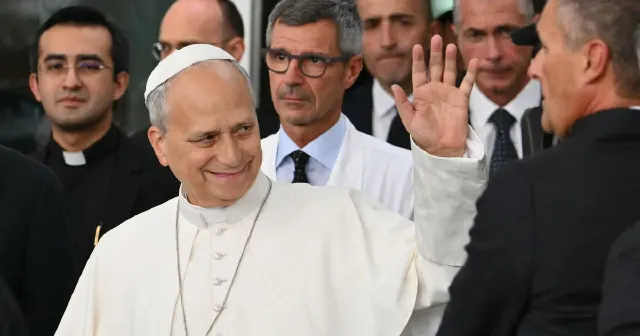 Elderly man in white papal-style vestments smiling and waving, surrounded by clergy and staff.