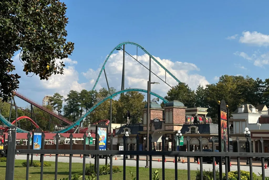Teal steel roller coaster track towering over an amusement park with fence and buildings