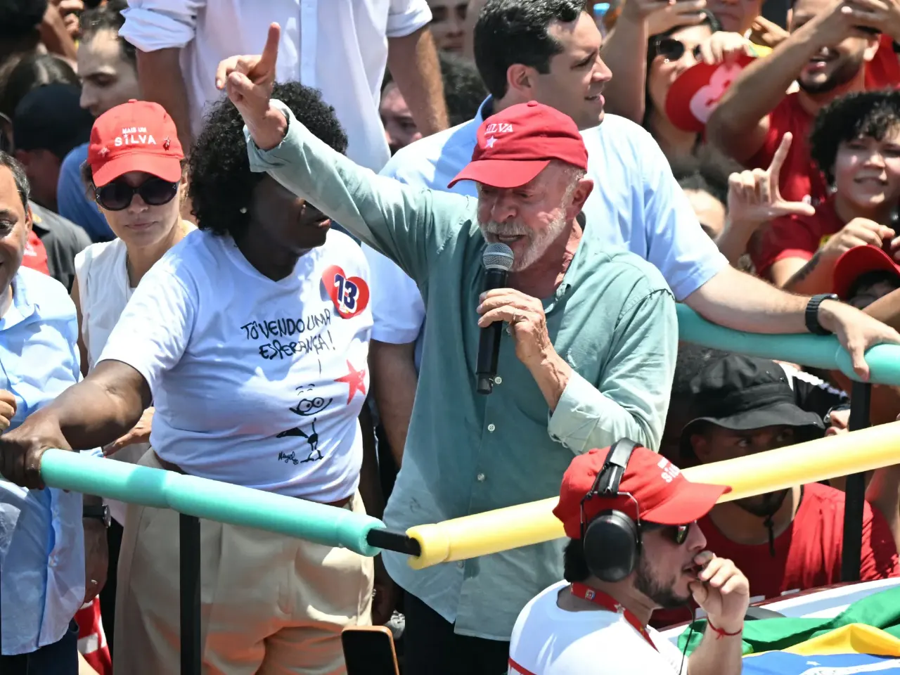 Older man in red cap speaks into microphone at outdoor rally, crowd in red caps and shirts behind him