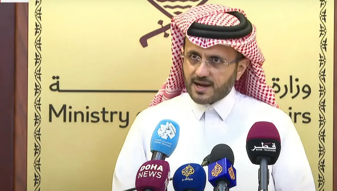 Man in white robe and red checkered headscarf speaking at press microphones before a Ministry of Foreign Affairs sign.