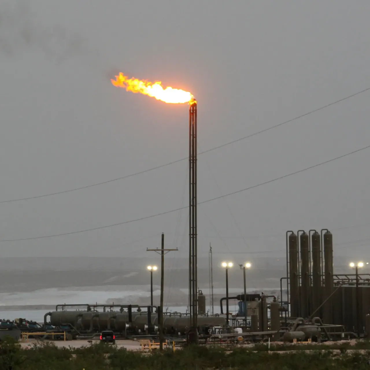 Gas flare burning atop a tall stack at an oil and gas facility at dusk.