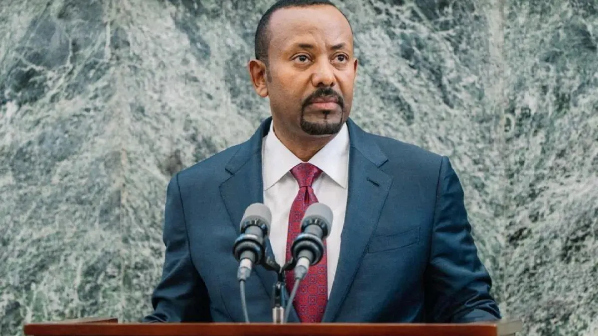 Man in dark suit and red tie speaking at a podium with microphones before green marble wall