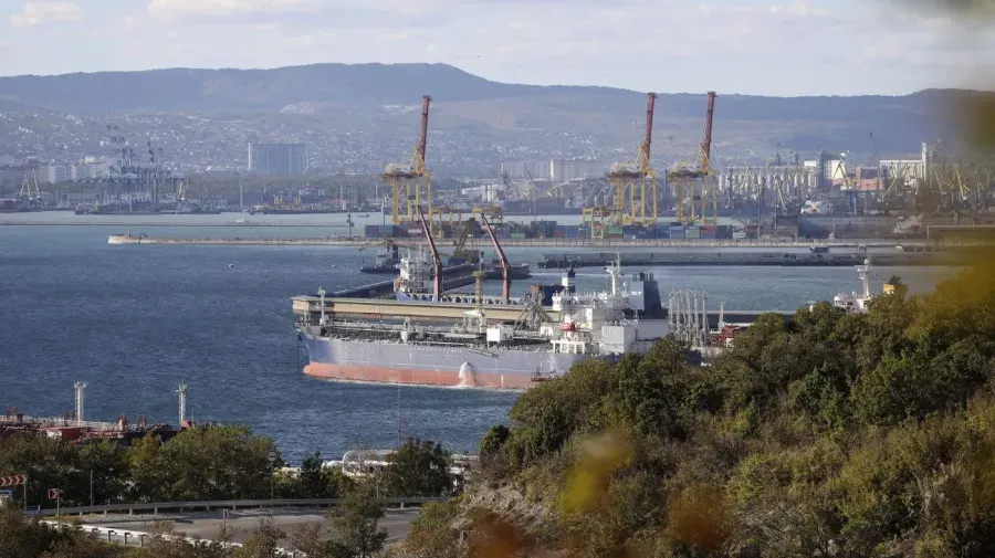 Large tanker ship docked at a port beside cranes, with hills and city behind.
