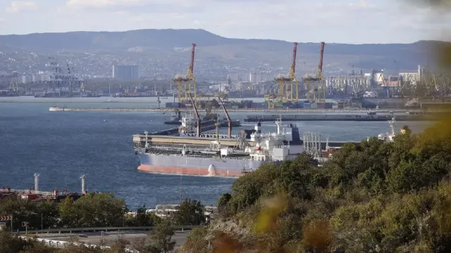 Large tanker ship docked at a port beside cranes, with hills and city behind.