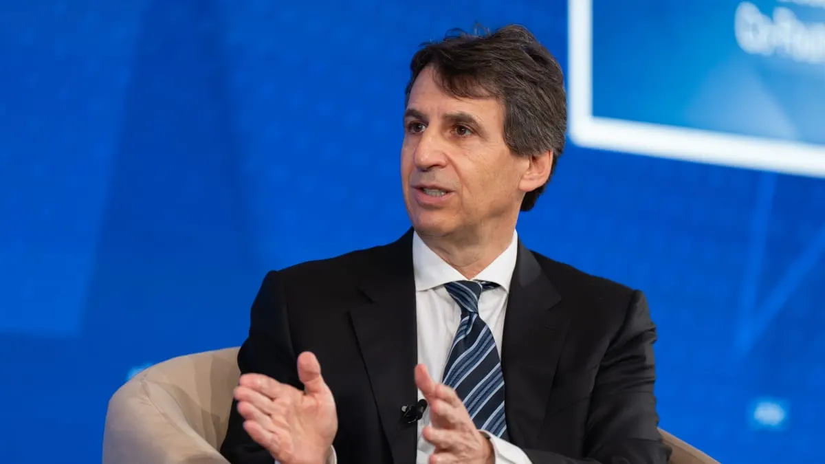 Man in dark suit and striped tie gesturing while speaking at a blue-backdrop conference panel.