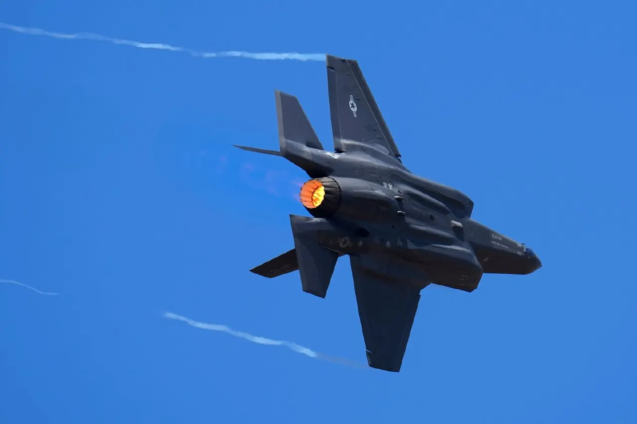 A dark F-35 fighter jet with glowing afterburner flying against a clear blue sky.