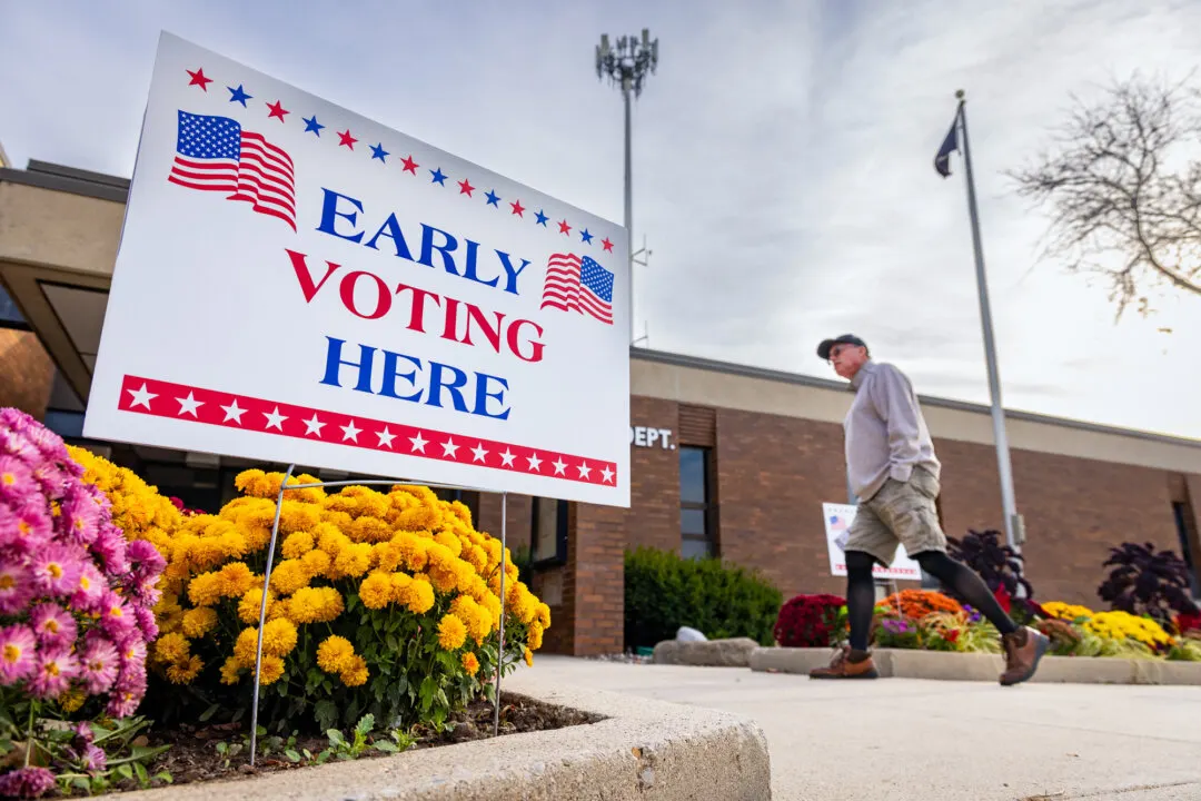Early Voting Here sign with American flags beside marigolds, with a man walking past a brick building.