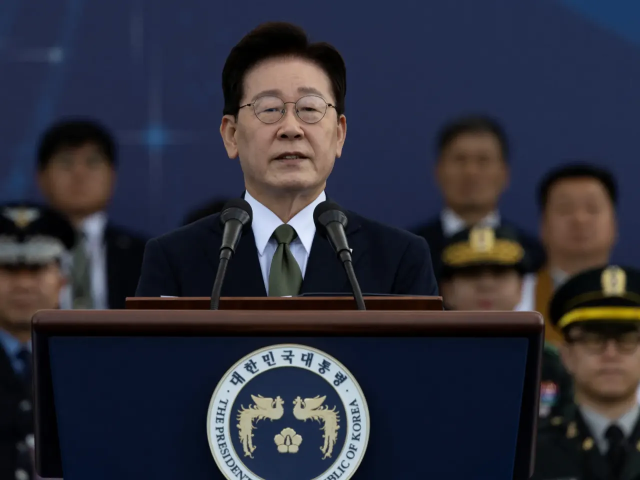 Man in dark suit speaking at podium bearing Presidential seal of Republic of Korea, with uniformed officials behind.
