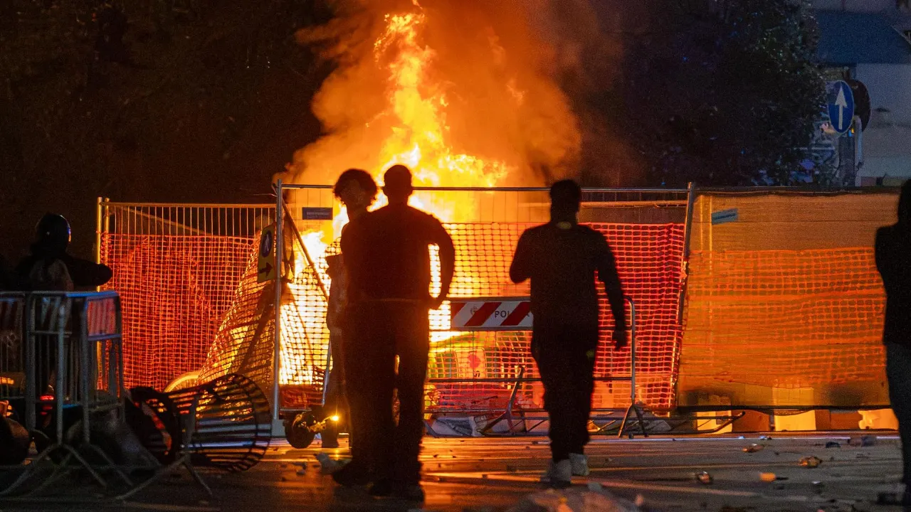 Silhouetted people watch a large fire burning behind orange barrier fencing at night, with debris on the street.