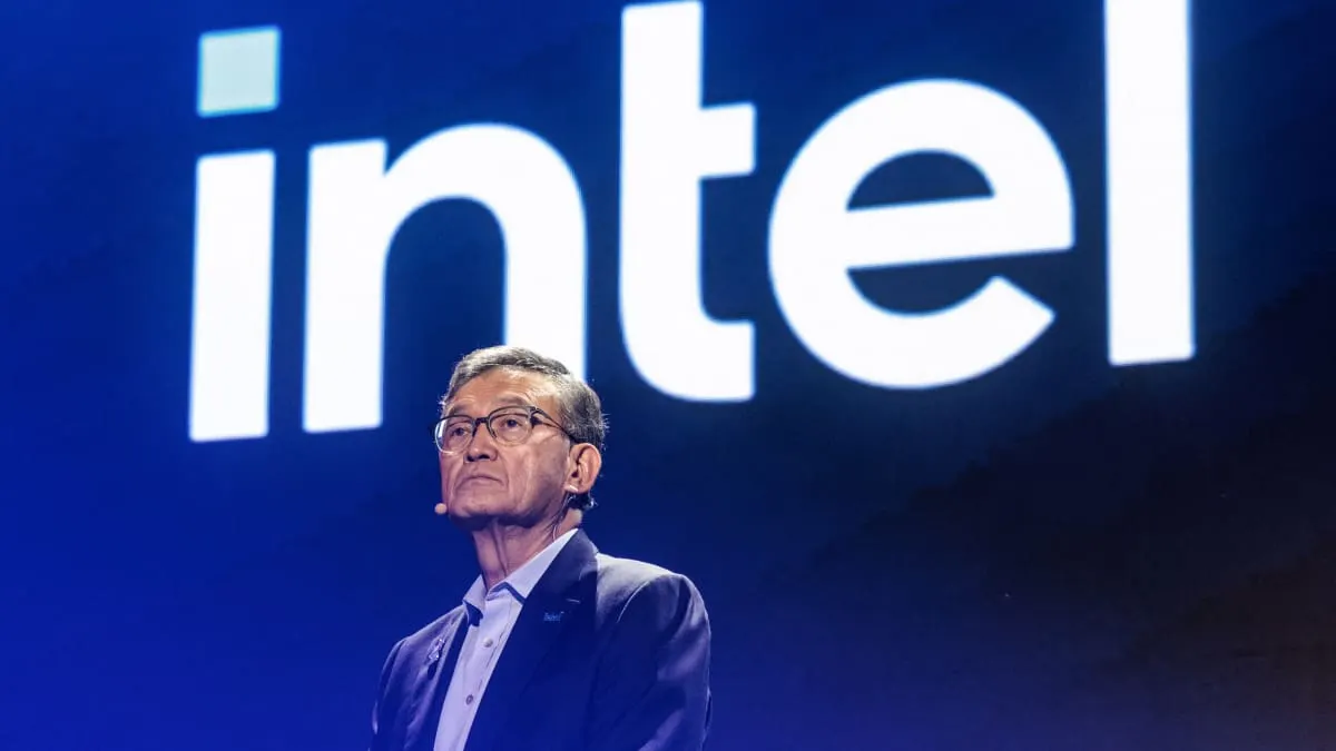Man in glasses and dark blazer on stage before large illuminated Intel logo.