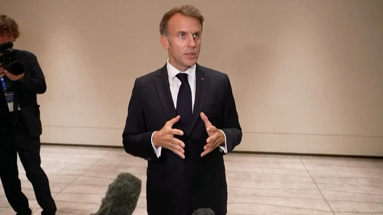 Man in dark suit gesturing while speaking to reporters, with cameraman and microphones visible.
