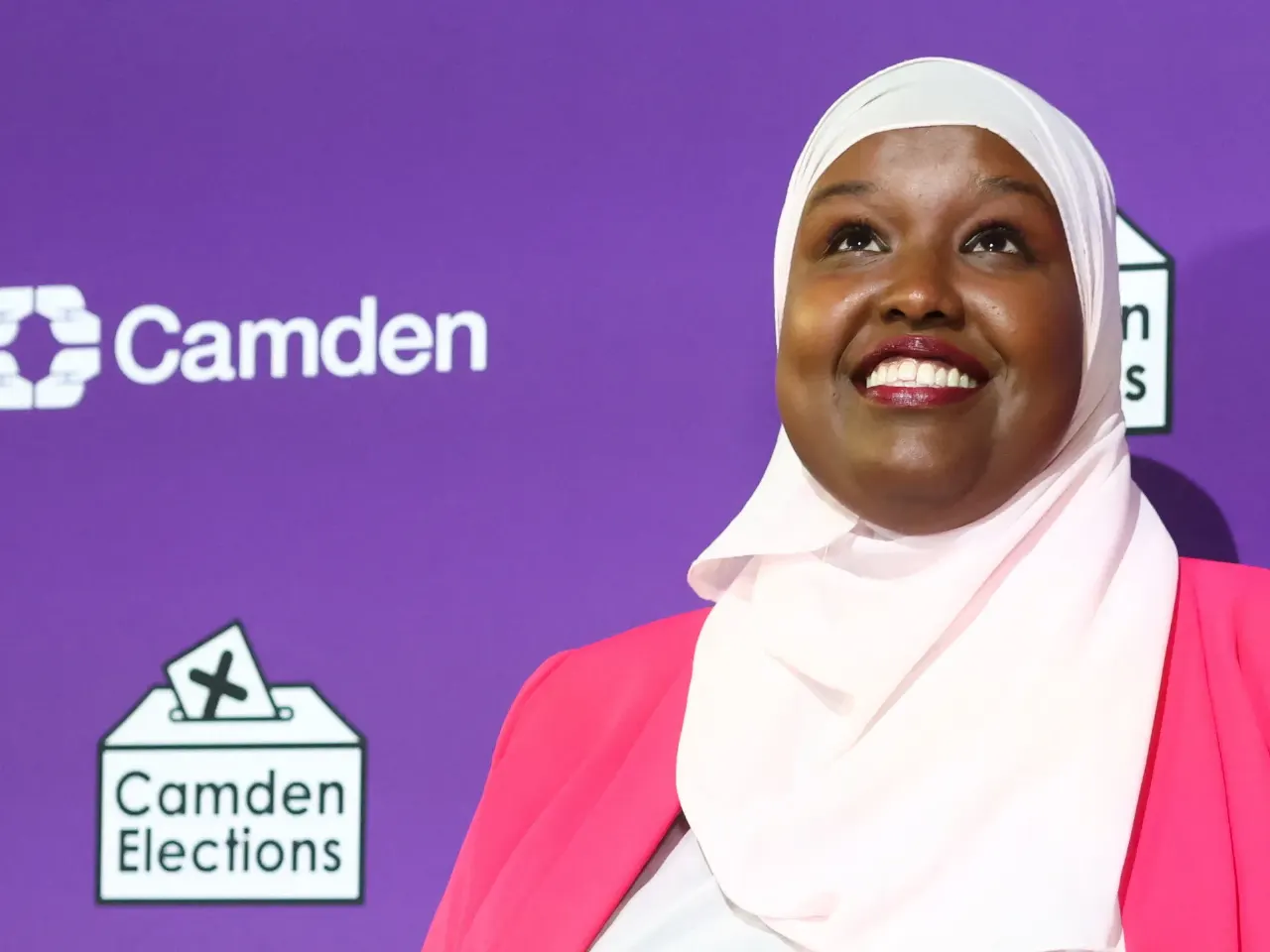 Smiling woman in white headscarf and pink jacket before purple Camden Elections backdrop.