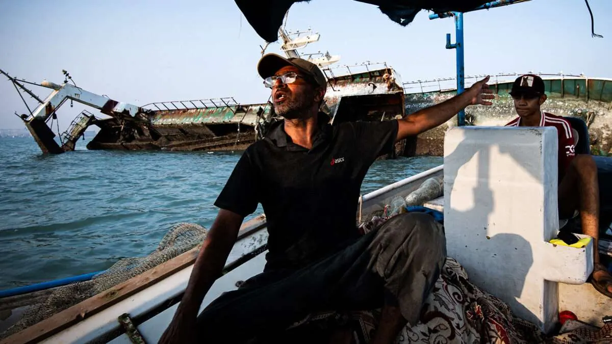 Man in cap gestures from a small boat toward a capsized ship in calm water.