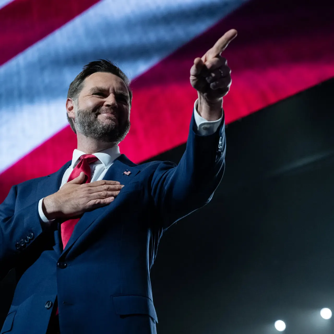 Man in navy suit and red tie pointing upward, hand on chest, before large American flag backdrop.