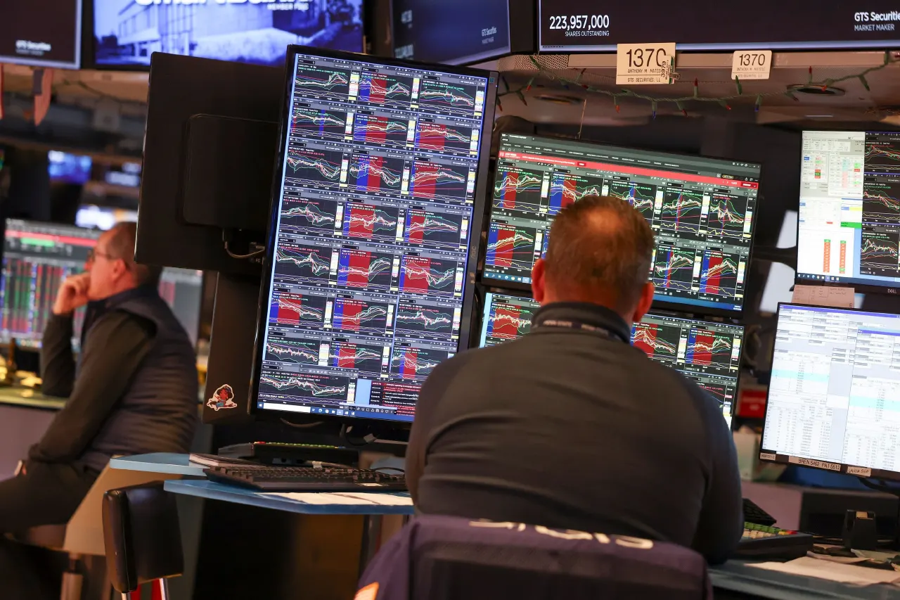 Traders at trading desks watching stock price charts on multiple monitors in a trading floor.